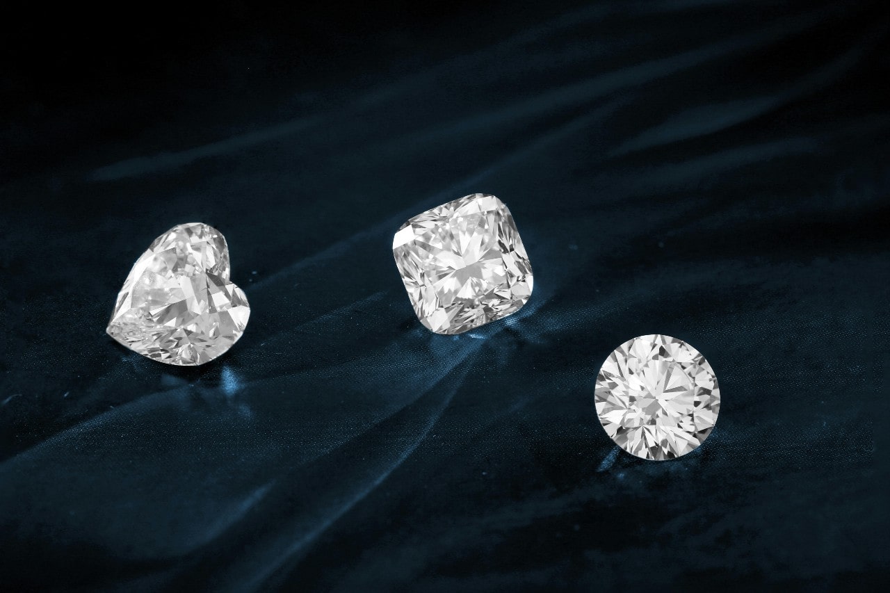 Three sparkling diamonds—heart, cushion, and round cuts—gleam on a dark, luxurious velvet fabric.