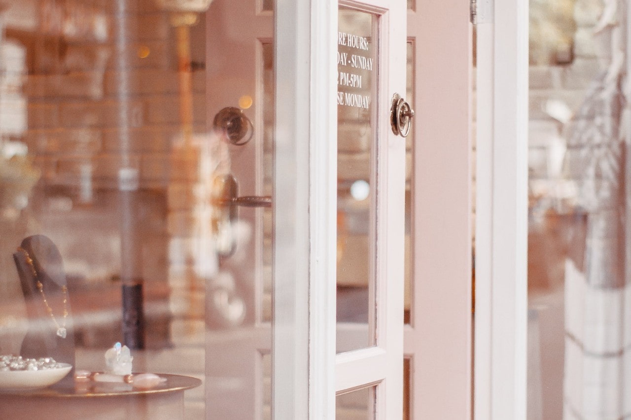 A view through a shop’s glass door showing jewelry displays and a reflection of the interior.