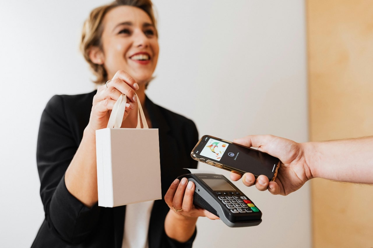A woman in a black blazer is holding a small white shopping bag watches as a person holds a smartphone over a contactless payment terminal.