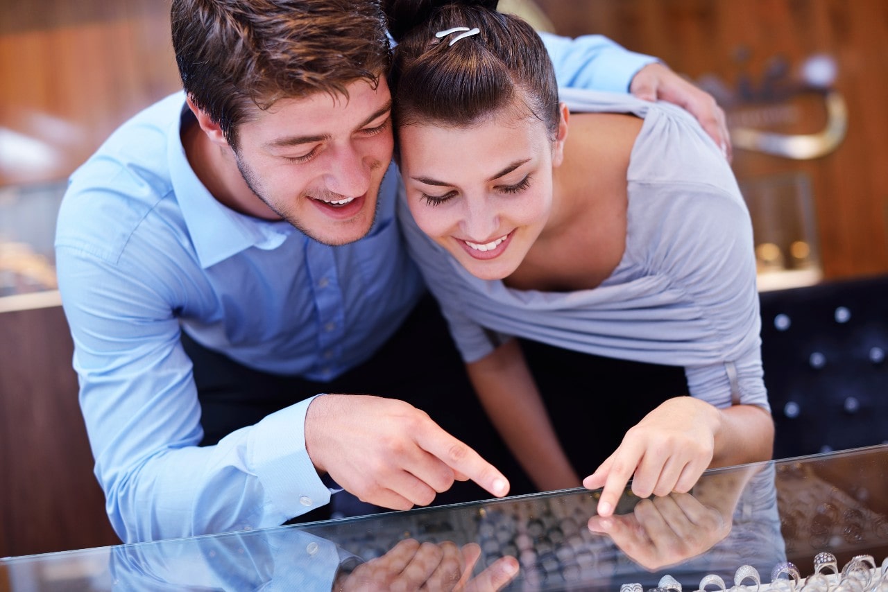 A smiling couple leans over a jewellery display case, excitedly pointing at rings inside.