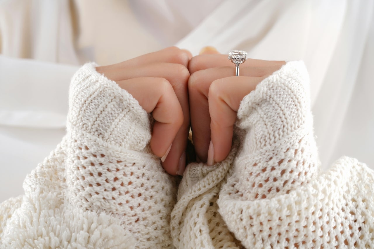 Close-up of a woman wearing a white knit sweater, showcasing a silver halo diamond engagement ring on her finger.