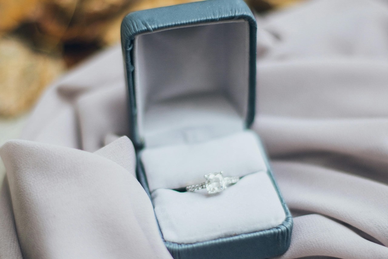 A white gold diamond side stone engagement ring in a white and mint velvet jewelry box on top of white fabric