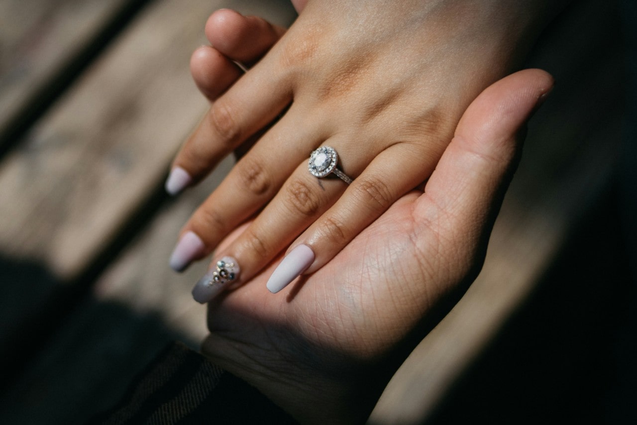 A woman’s hand with a long pink manicure is held by a man's hand, showcasing a halo oval engagement ring in a sunlight setting.
