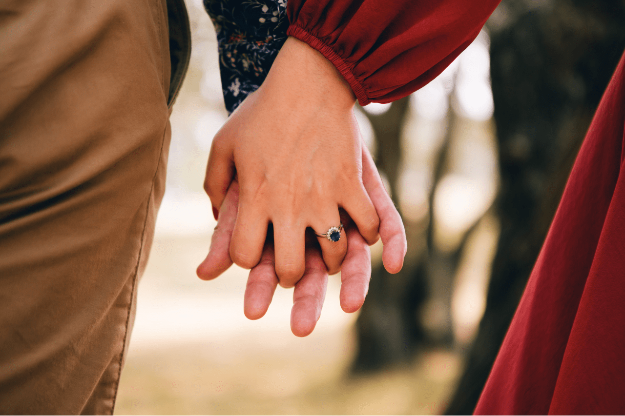 A sapphire engagement ring with a diamond halo on the hand of a woman holding hands with her partner.