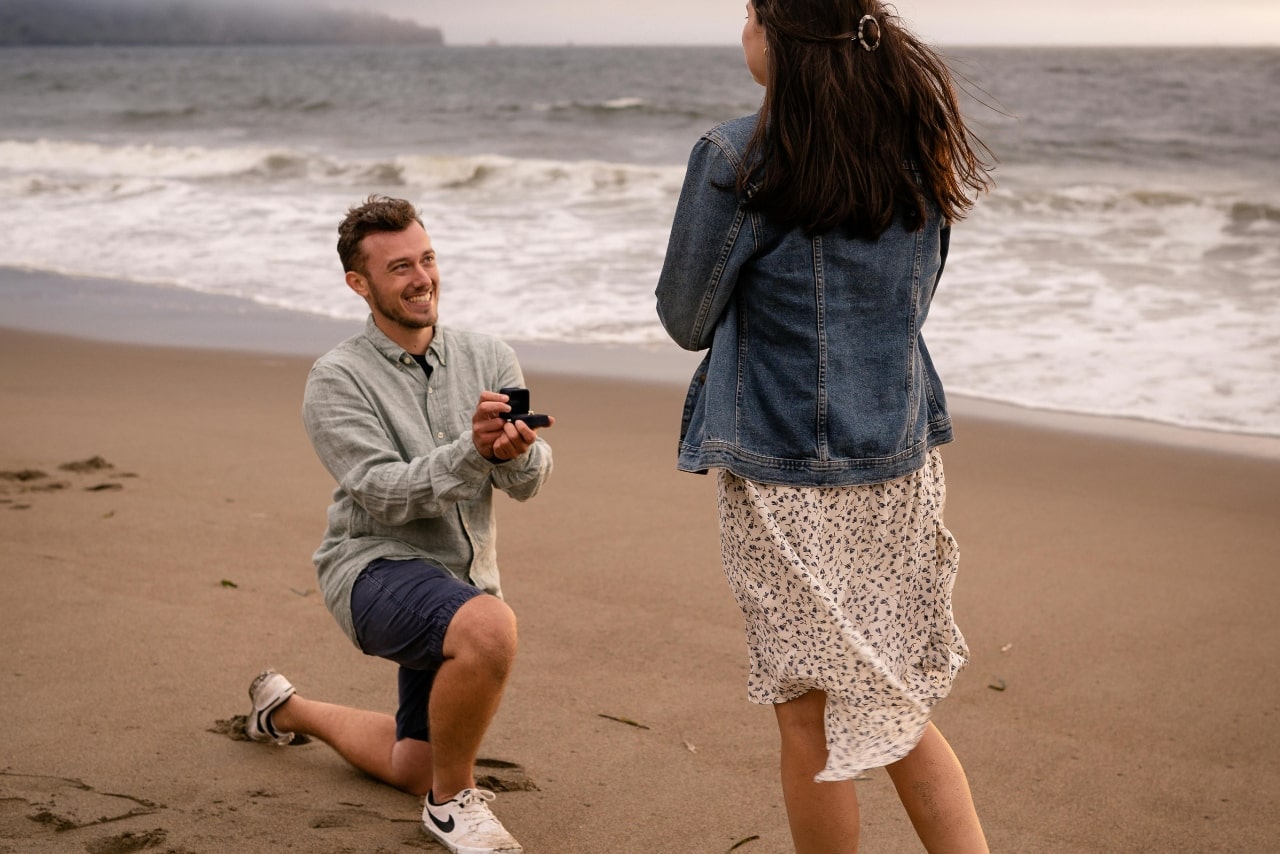 A man proposing to a woman in a dress on a beach.