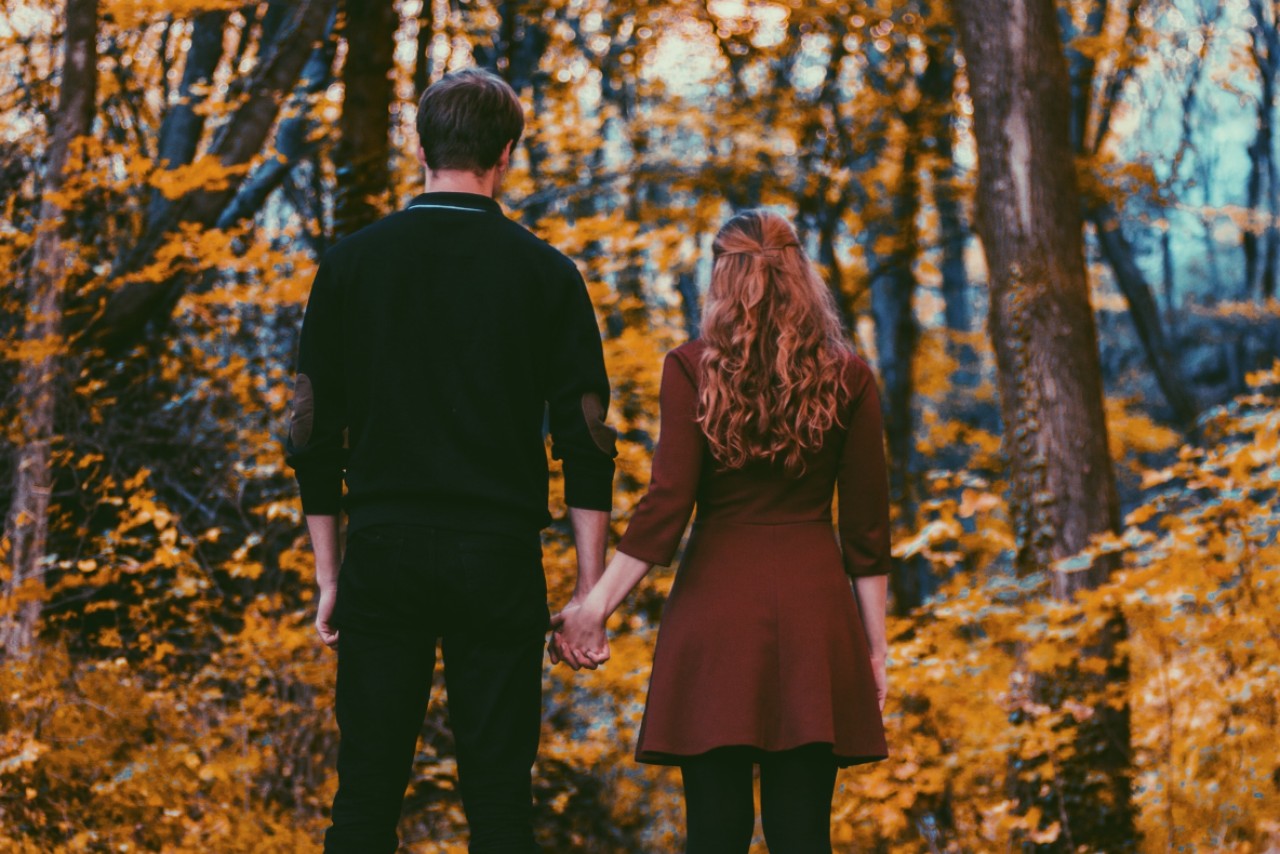 A couple stands hand-in-hand in front of an autumn forest, surrounded by orange leaves.