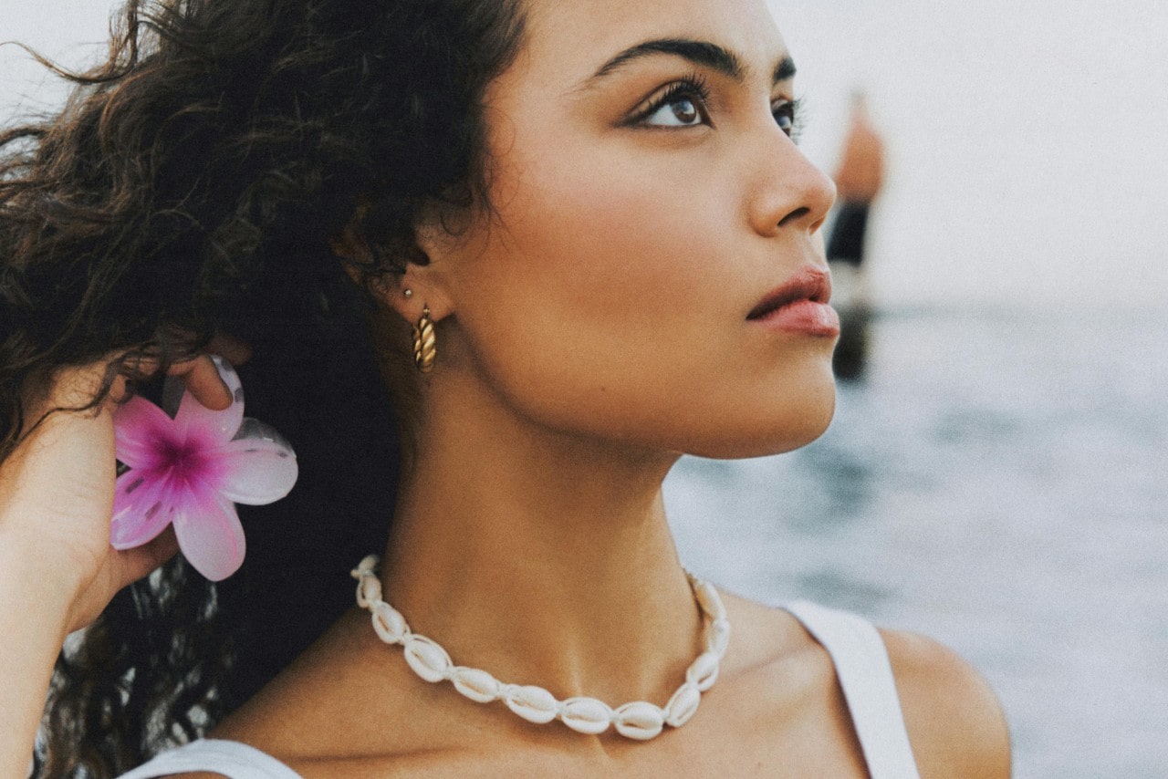 A close-up of a woman on the beach wearing a cowrie shell necklace and holding a flower.