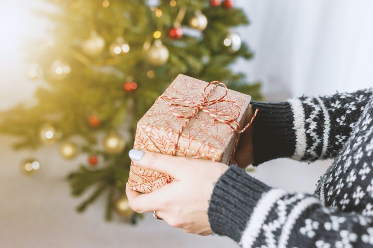 A close up of wrapped gift in a hands in gray sweater against a blurred christmas tree