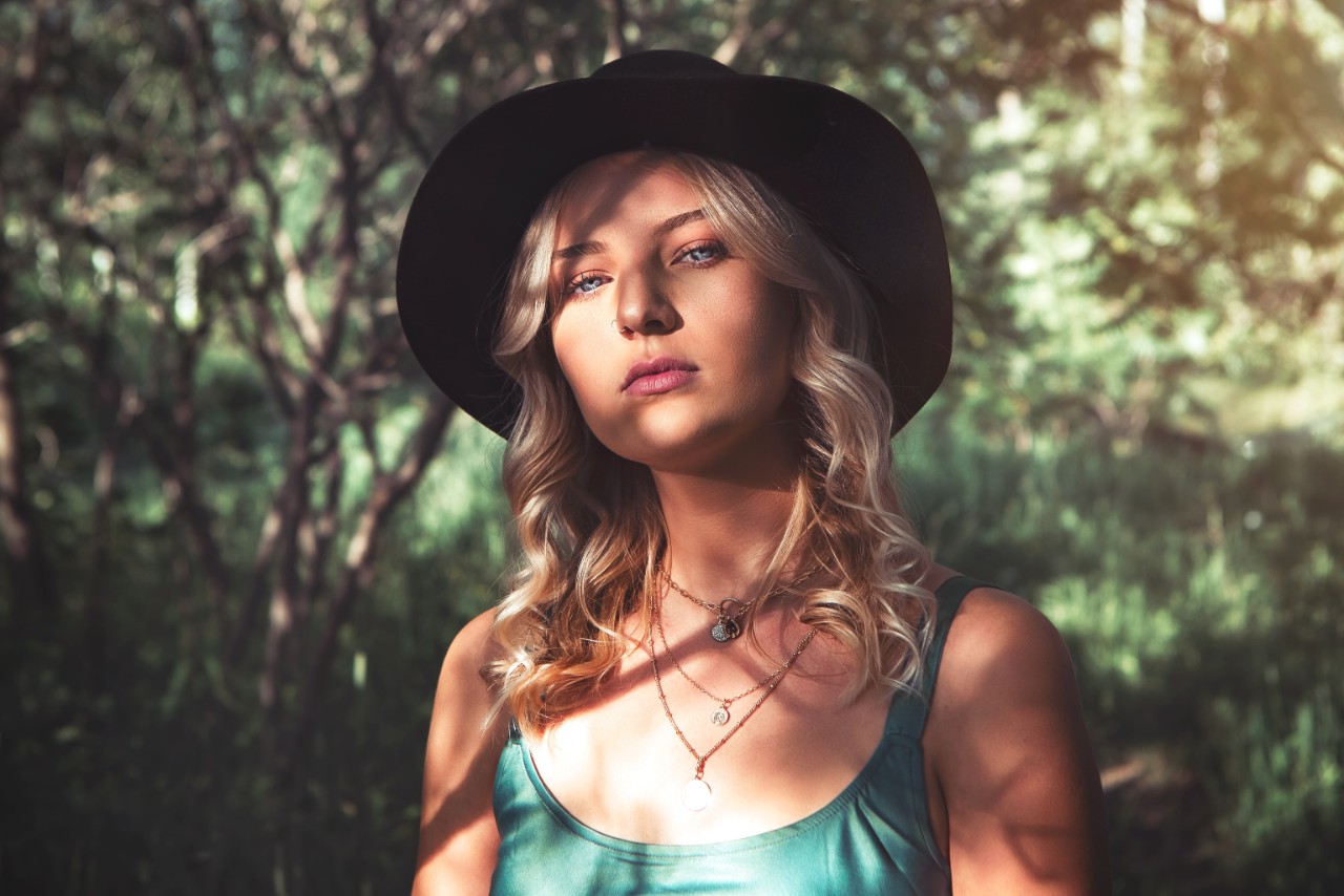 A young blond woman in a hat and green top stands in front of the forest, showcasing layered pendant necklaces.