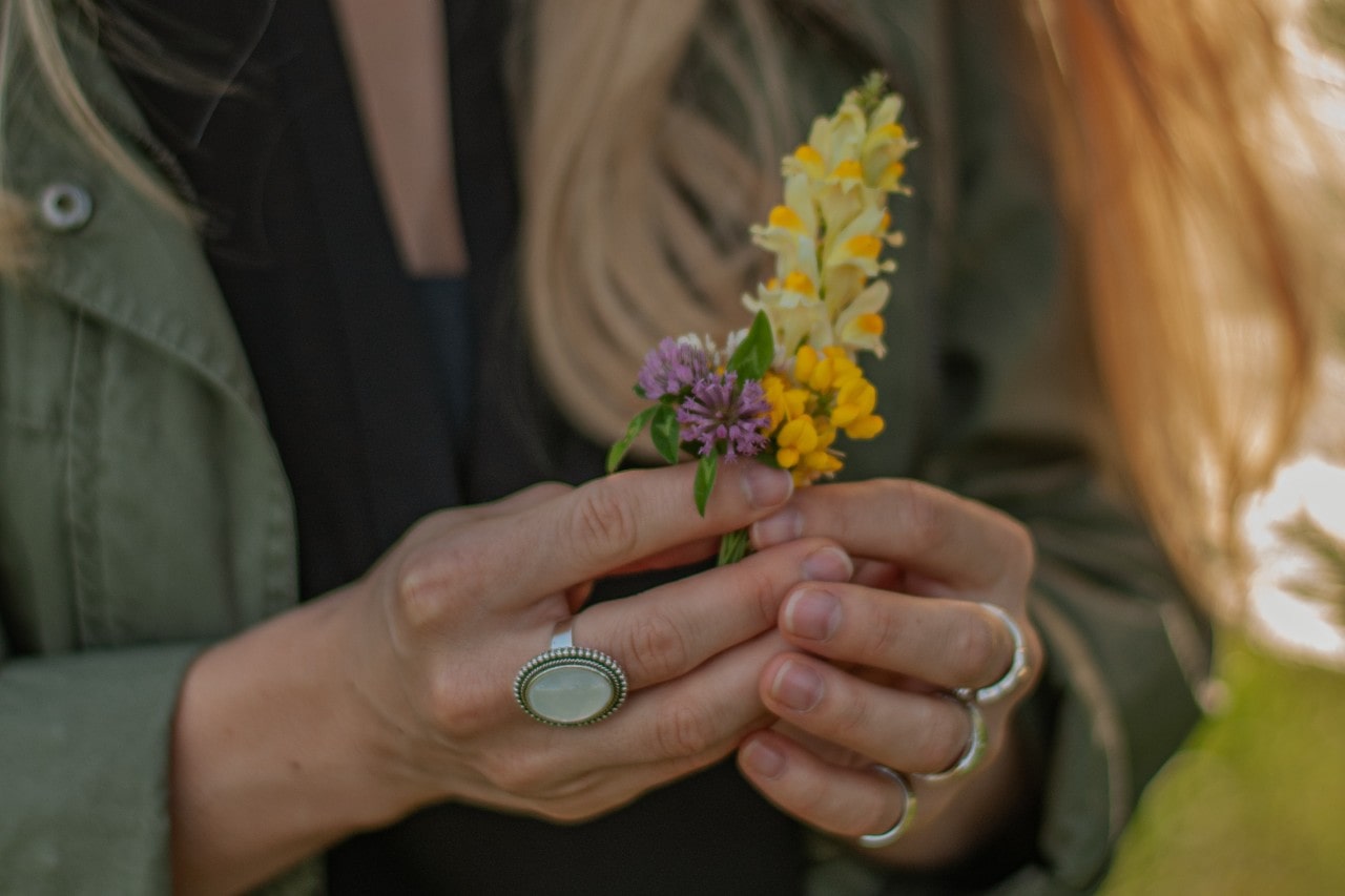 A close up of a woman&rsquo;s hands adorned with silver rings holding yellow and purple flowers.