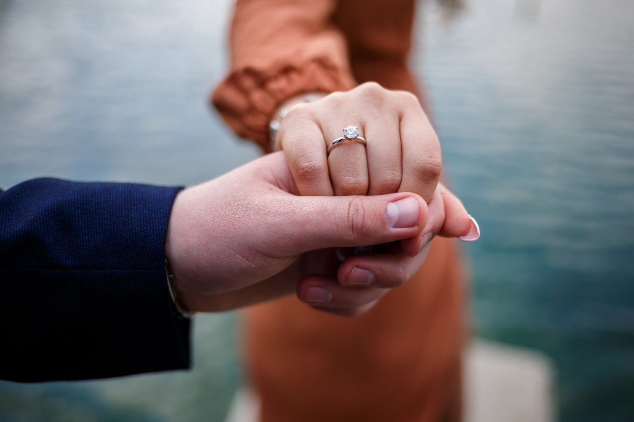 Close-up of two hands holding by a serene waterfront, one adorned with a sparkling engagement ring