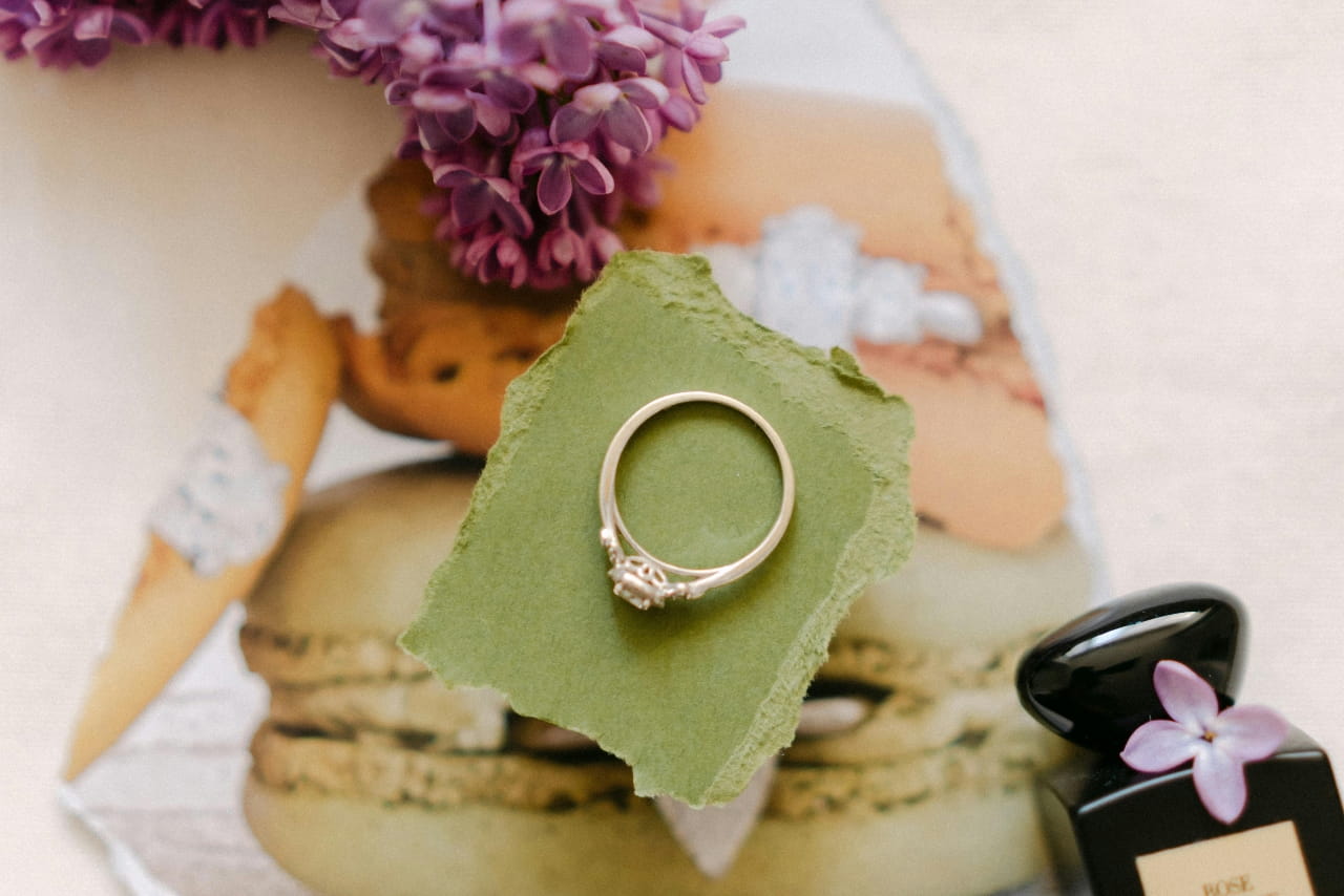 A close up of an engagement ring with a cathedral setting on top of a green piece of paper with flowers and a picture of macarons in the background.