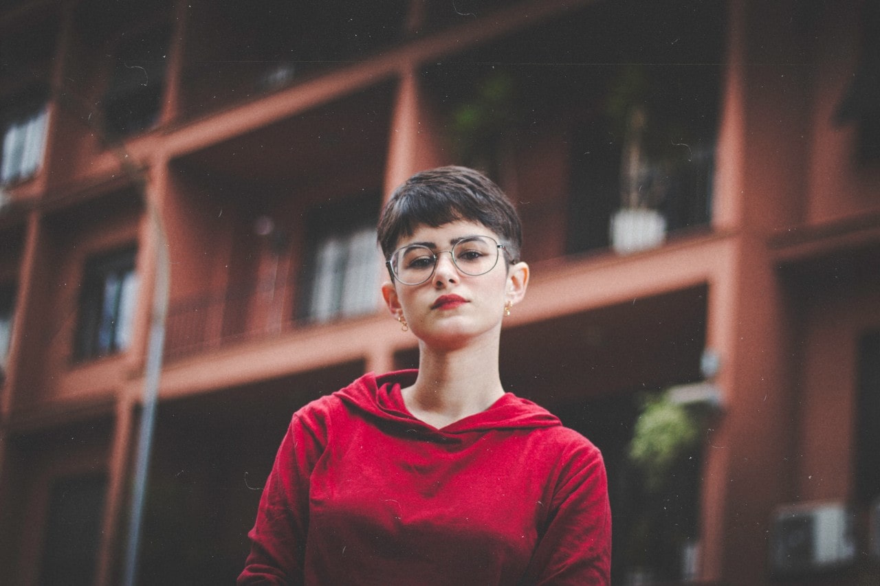 A short-haired woman in red wears gold hoop earrings outside an apartment complex.