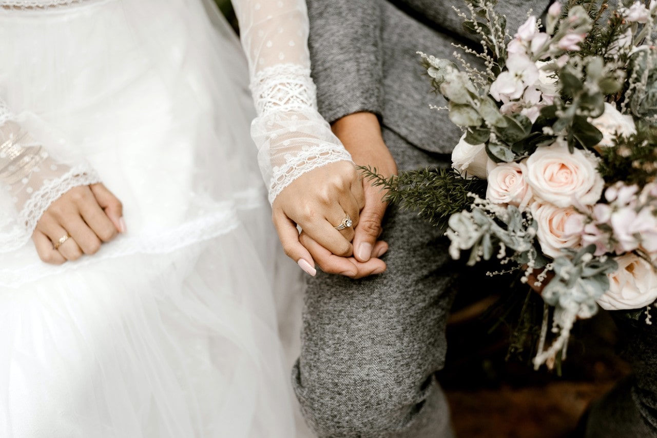 Bride in white and groom in grey sharing a quiet moment as her diamond ring catches the light with a bouquet in his hand.