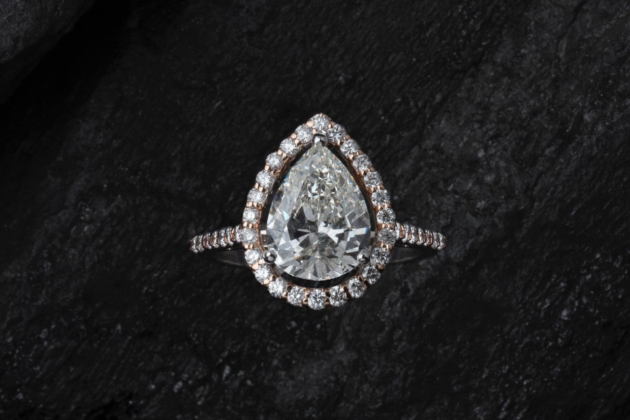 A close up of pear-shaped diamond ring with center stone and side accent diamonds resting on black background