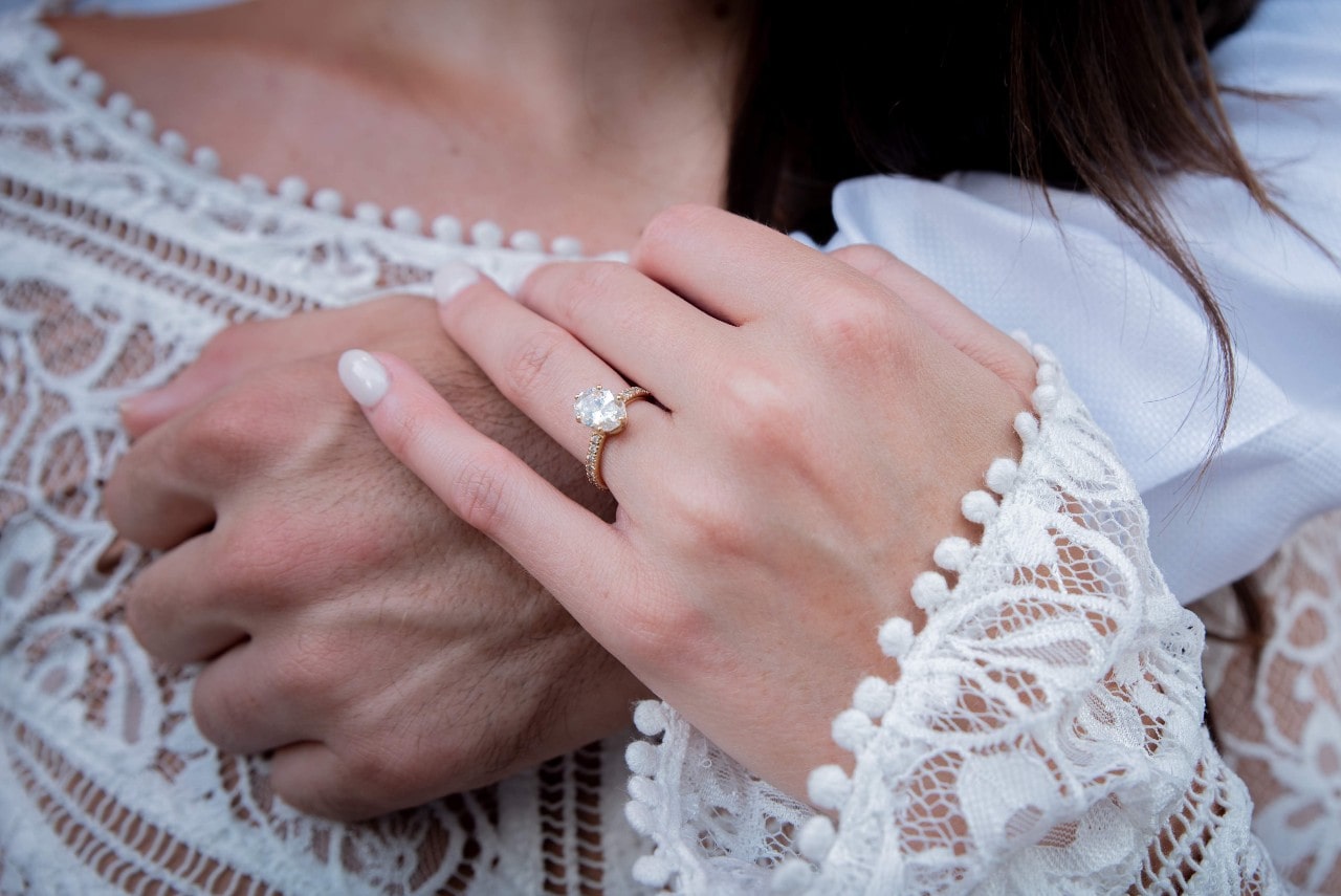 A close-up of a woman showcasing her oval diamond engagement ring as she holds man’s hand to her heart, while wearing white blouse.