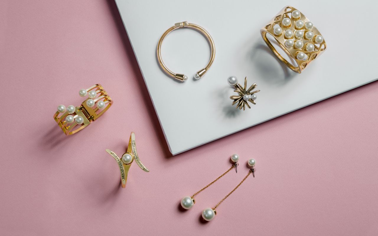 A collection of elegant gold jewelry with pearls on a pink background. Includes bracelets, a ring, and dangling earrings.