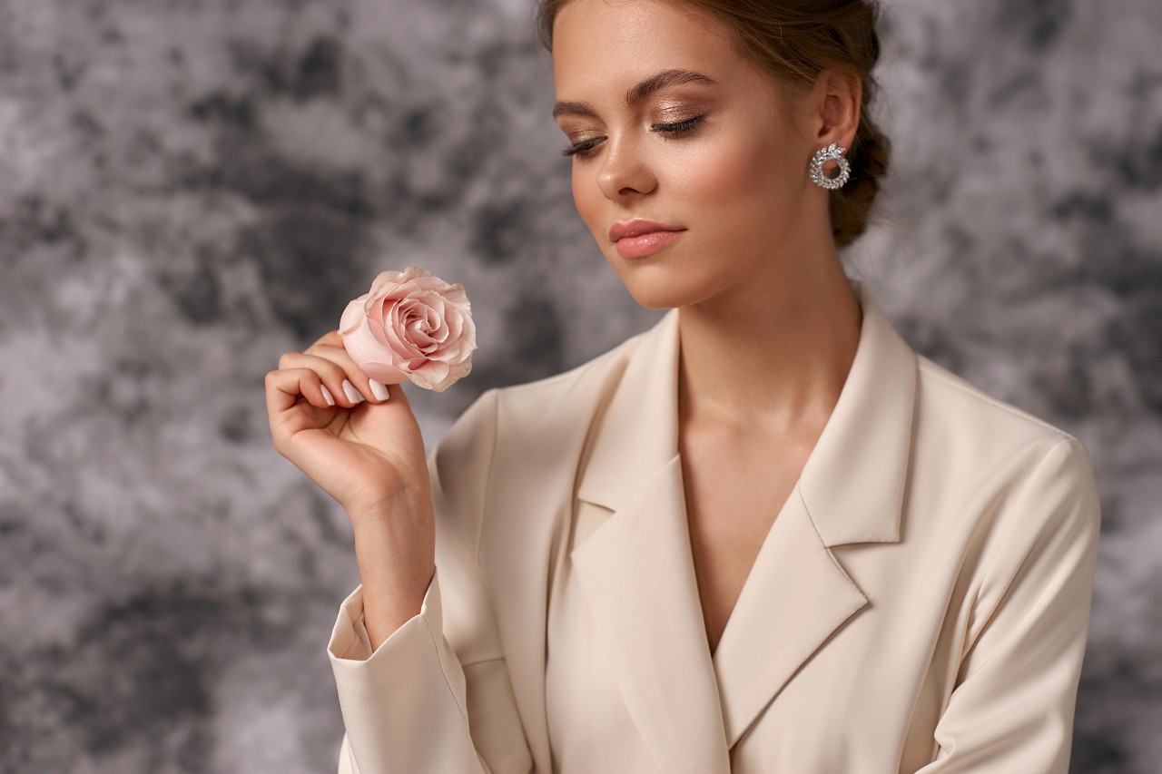 A woman in a beige blaze holds a pink rose while wearing round diamond earrings. A woman in a beige blaze holds a pink rose while wearing round diamond earrings.