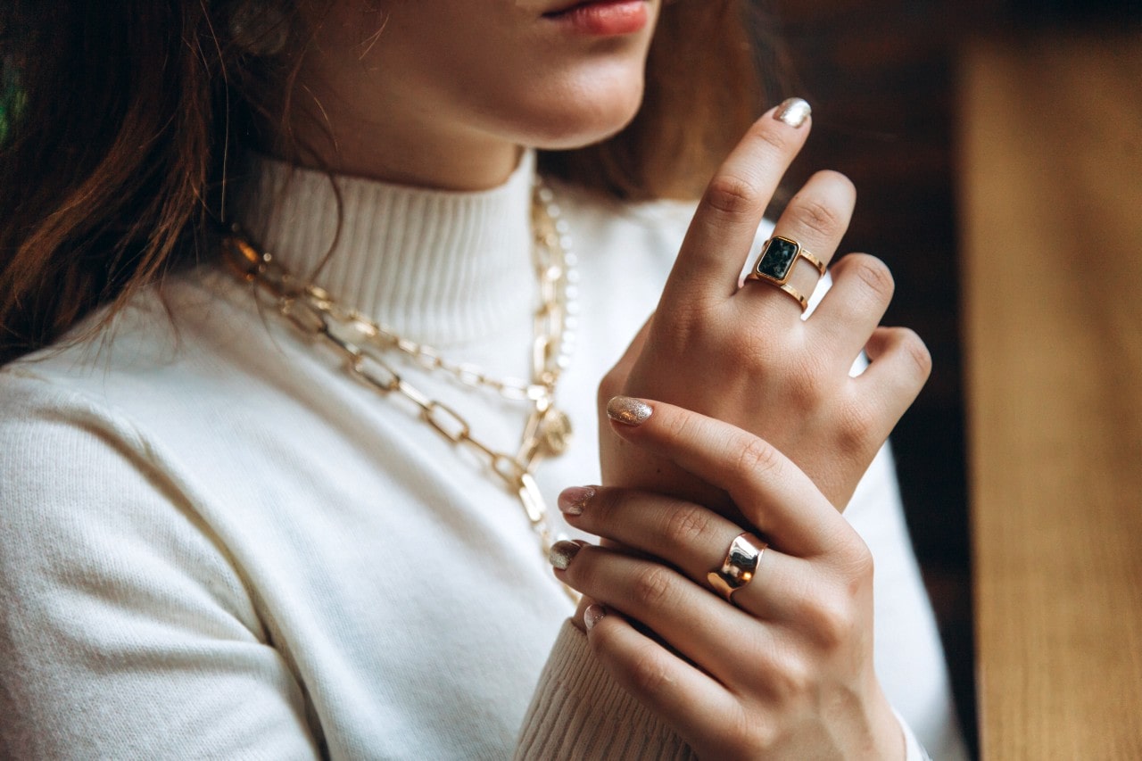 A woman in a white turtleneck showcases layered yellow gold chain necklaces and chunky fashion rings. A woman in a white turtleneck showcases layered yellow gold chain necklaces and chunky fashion rings.