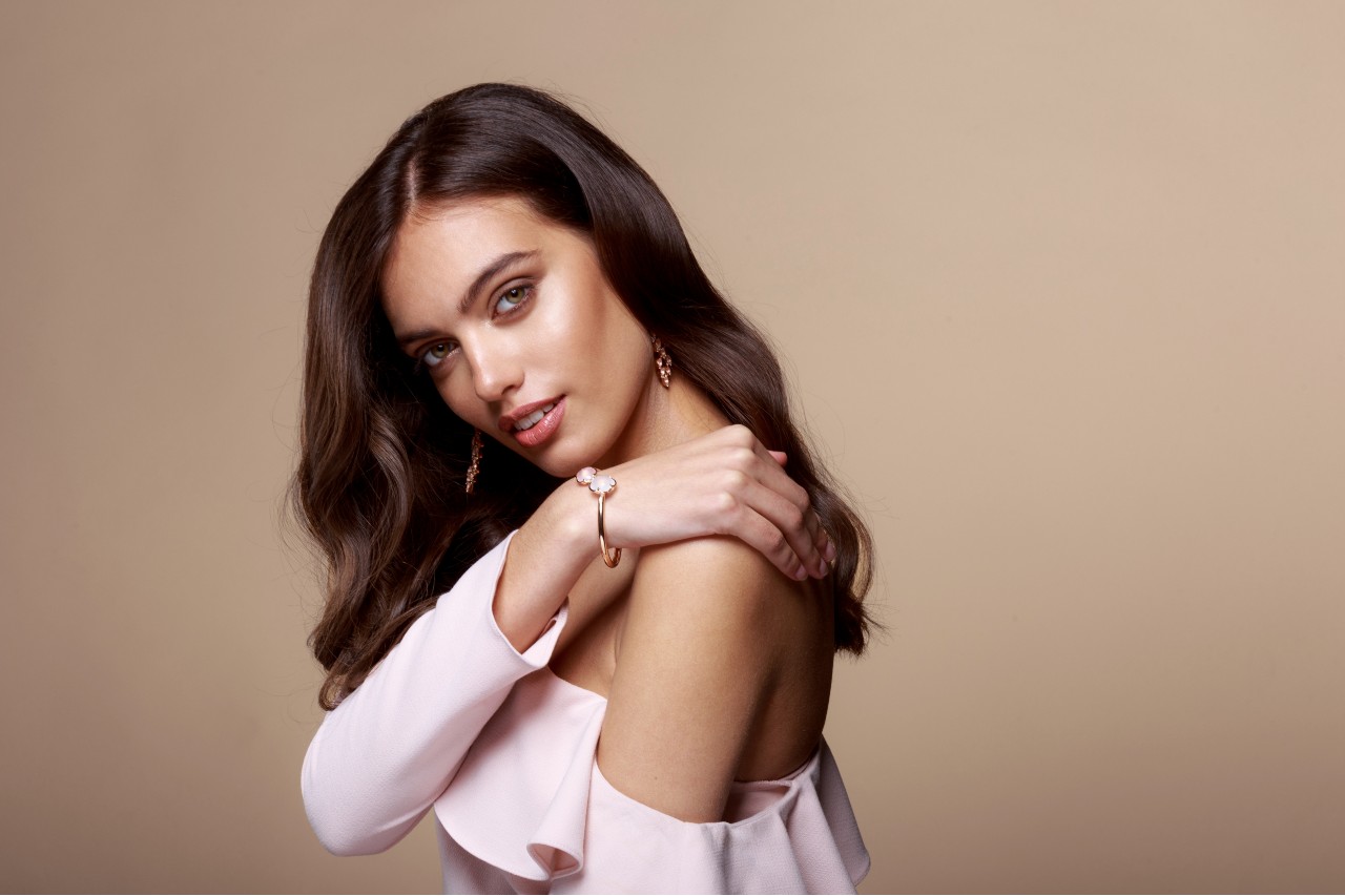 A woman with brunette hair in a pink blouse wears a rose gold floral bangle and matching drop earrings. A woman with brunette hair in a pink blouse wears a rose gold floral bangle and matching drop earrings.