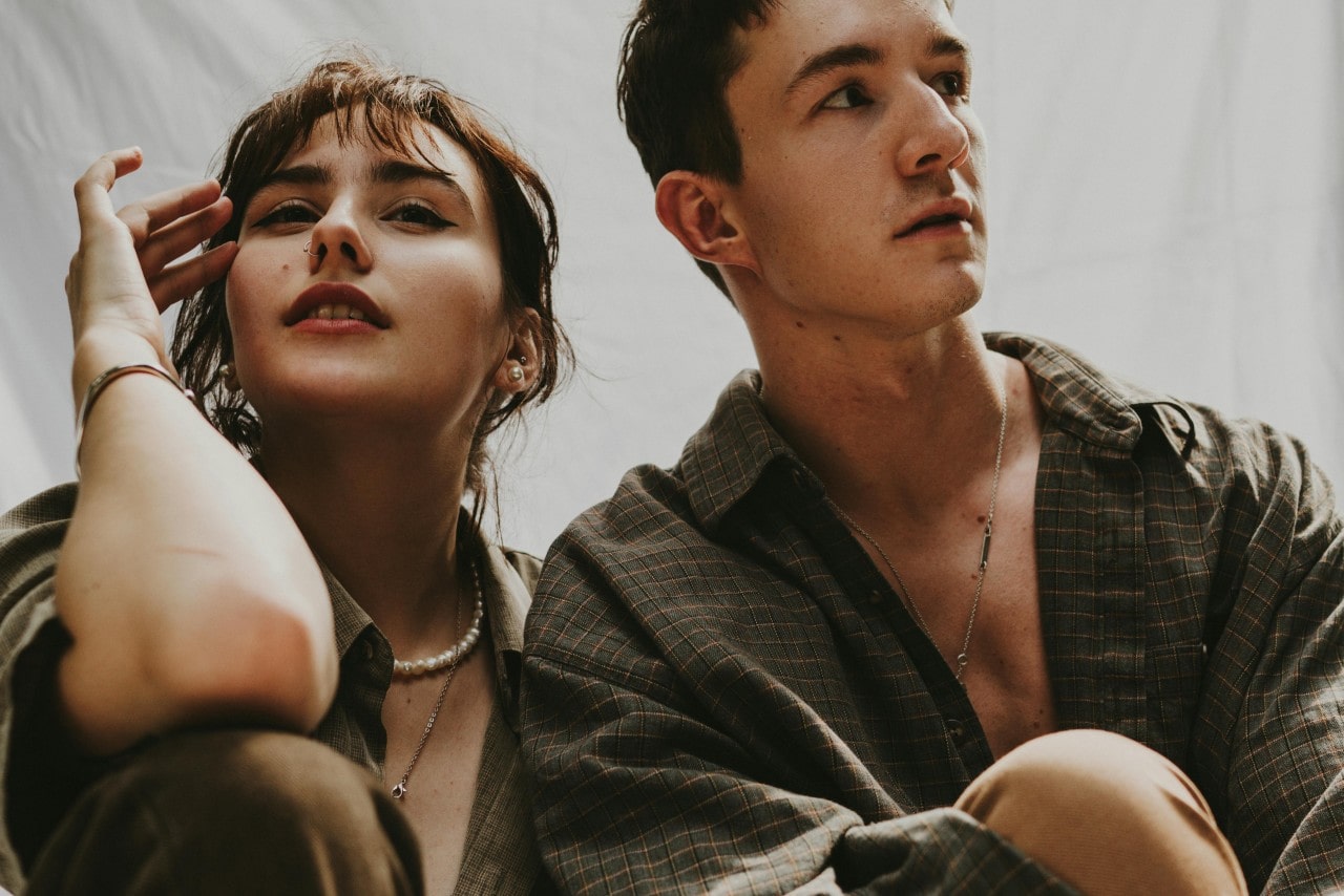 A close-up of a casually dressed couple, the man wearing a distinctive necklace and the woman wearing layered necklaces, a bangle bracelet, and stud earrings.