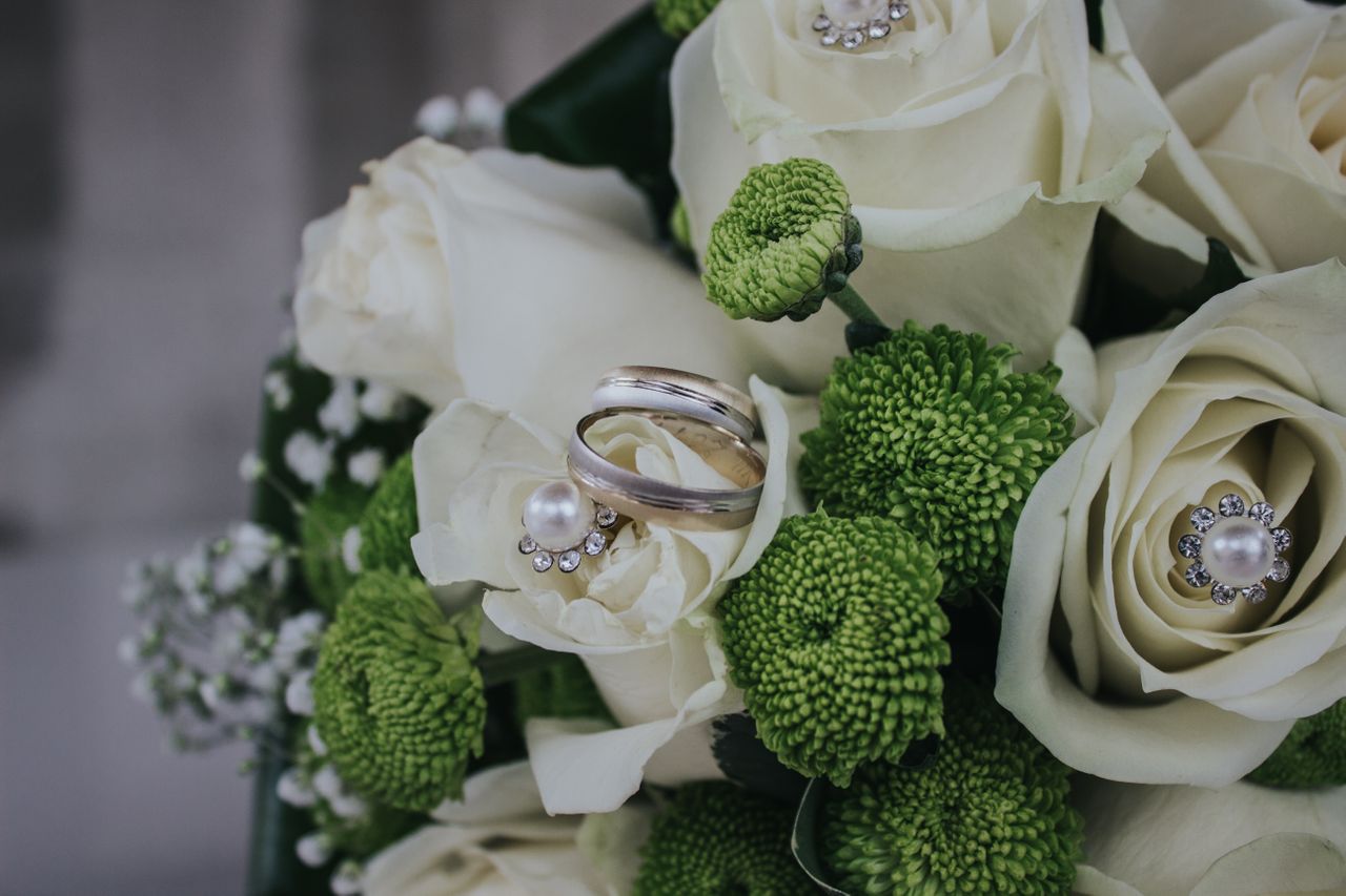 A pair of silver wedding rings rest on white roses adorned with pearls and diamonds, surrounded by green chrysanthemums. A pair of silver wedding rings rest on white roses adorned with pearls and diamonds, surrounded by green chrysanthemums.
