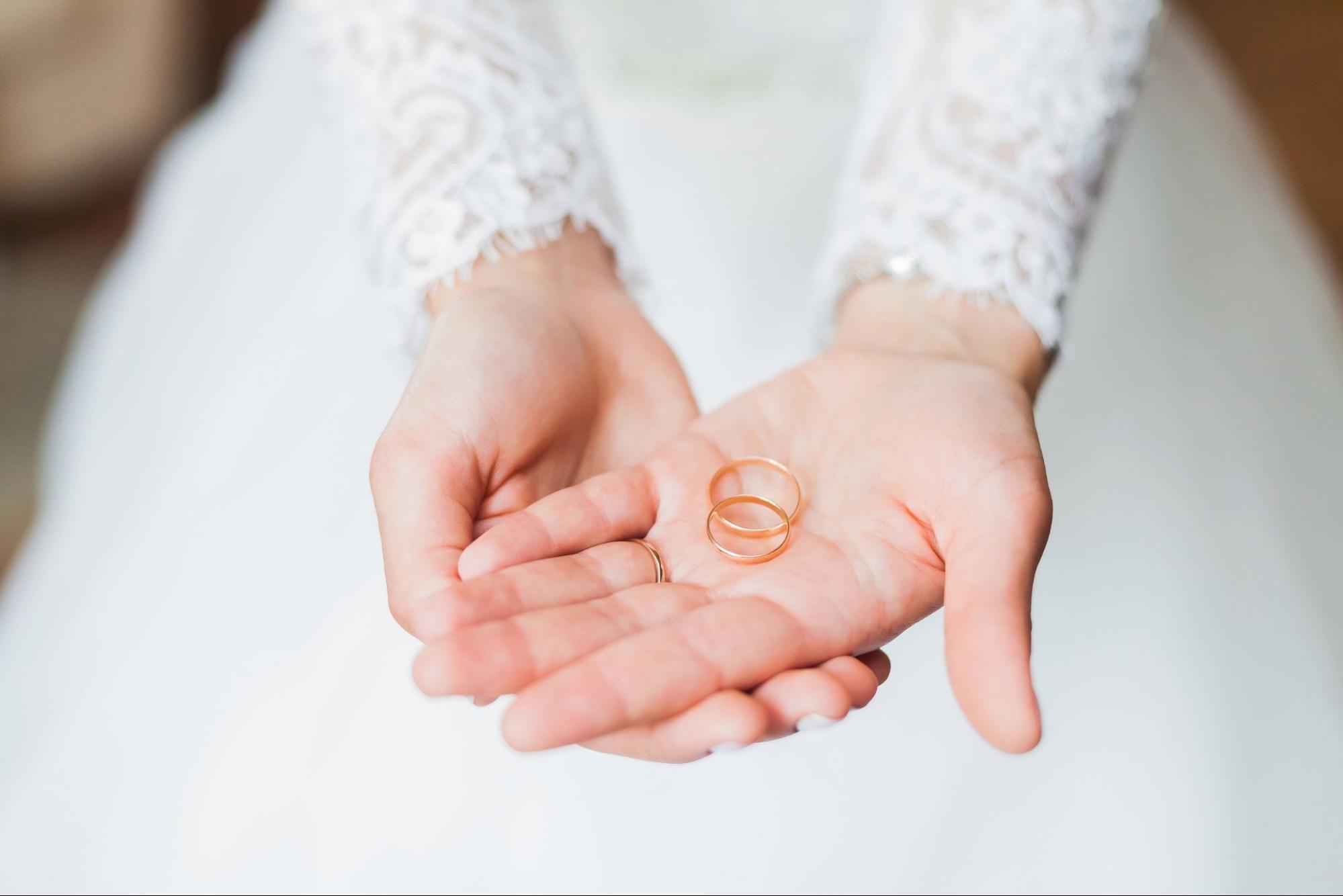 A close-up of a bride’s hands in a lace white gown, showcasing two thin yellow gold wedding bands on her palm. A close-up of a bride’s hands in a lace white gown, showcasing two thin yellow gold wedding bands on her palm.