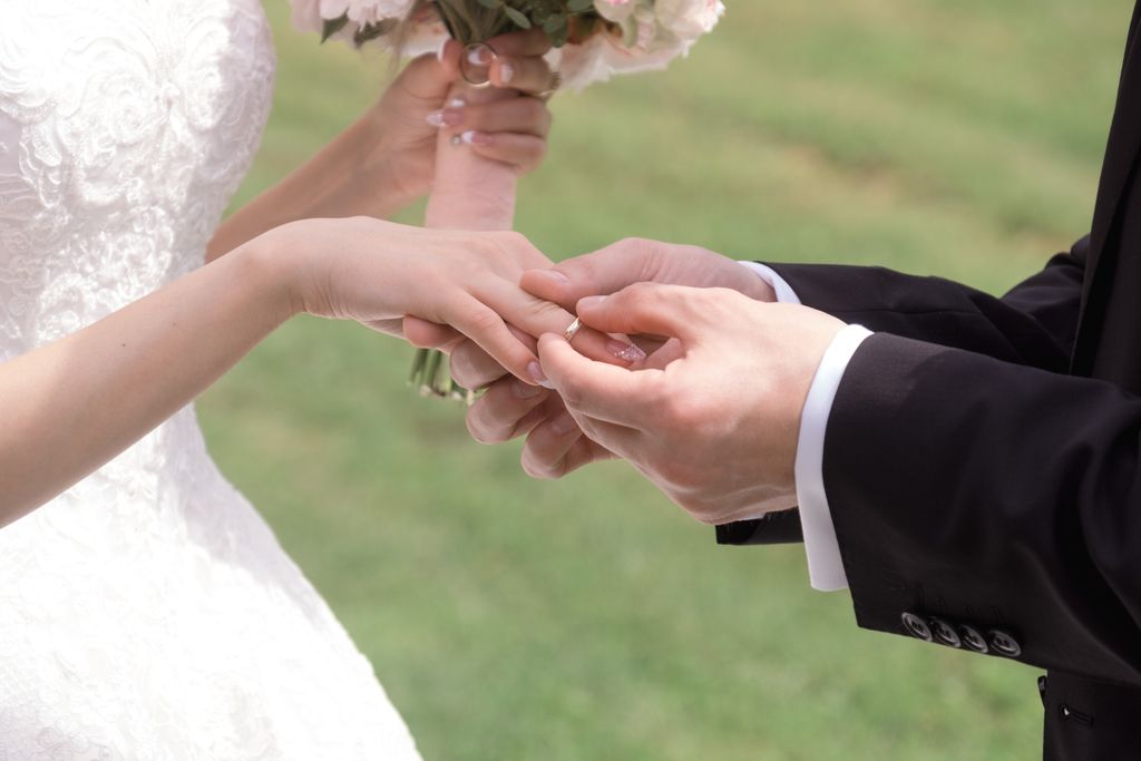 A groom puts a wedding band on a bride’s hand, while she is in a white dress holding a bouquet in the other hand, against a blurry grass. A groom puts a wedding band on a bride’s hand, while she is in a white dress holding a bouquet in the other hand, against a blurry grass.