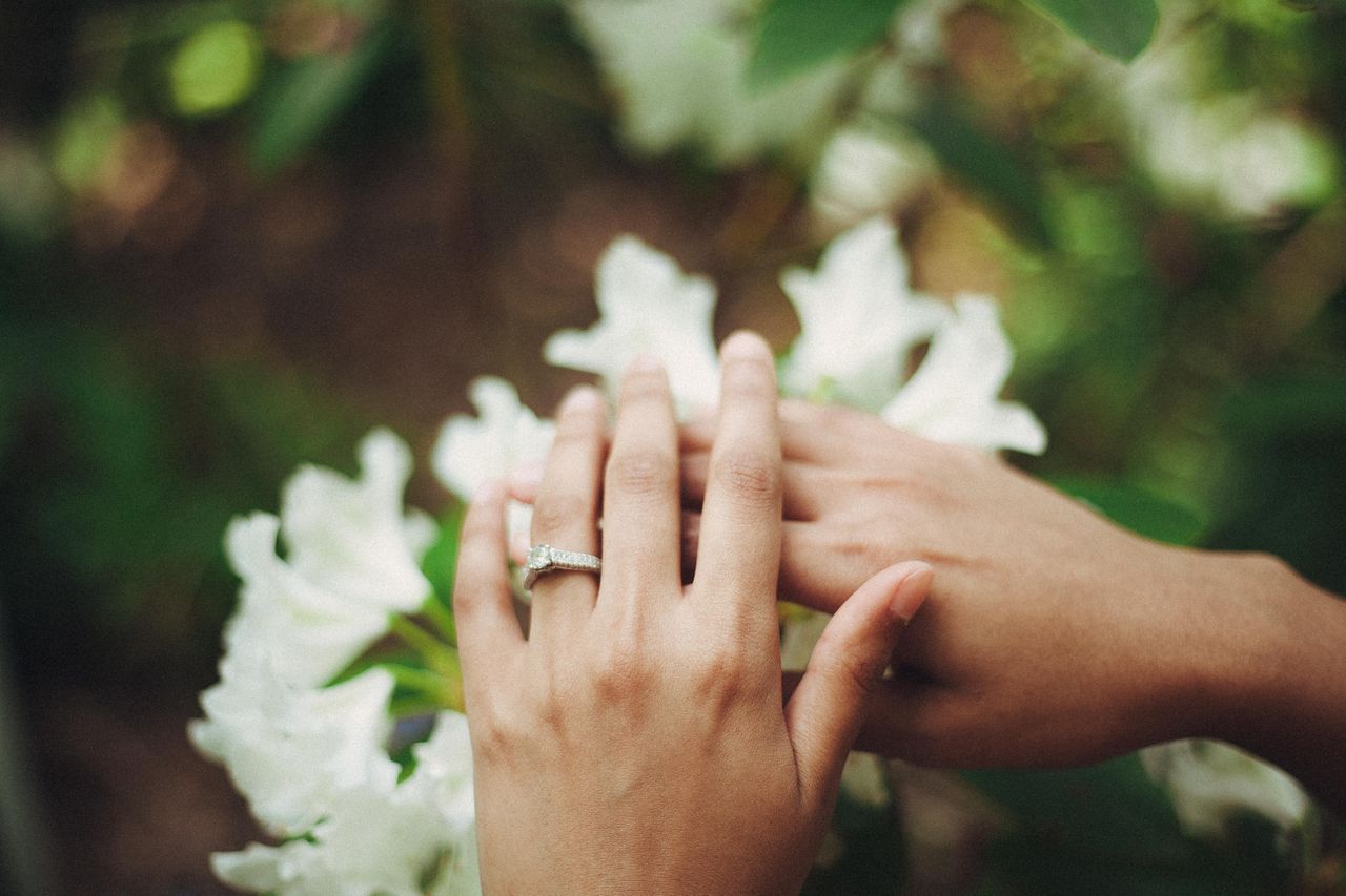 A pair of bride’s hands touching near white flowers, with a diamond engagement ring visible on the finger. A pair of bride’s hands touching near white flowers, with a diamond engagement ring visible on the finger.