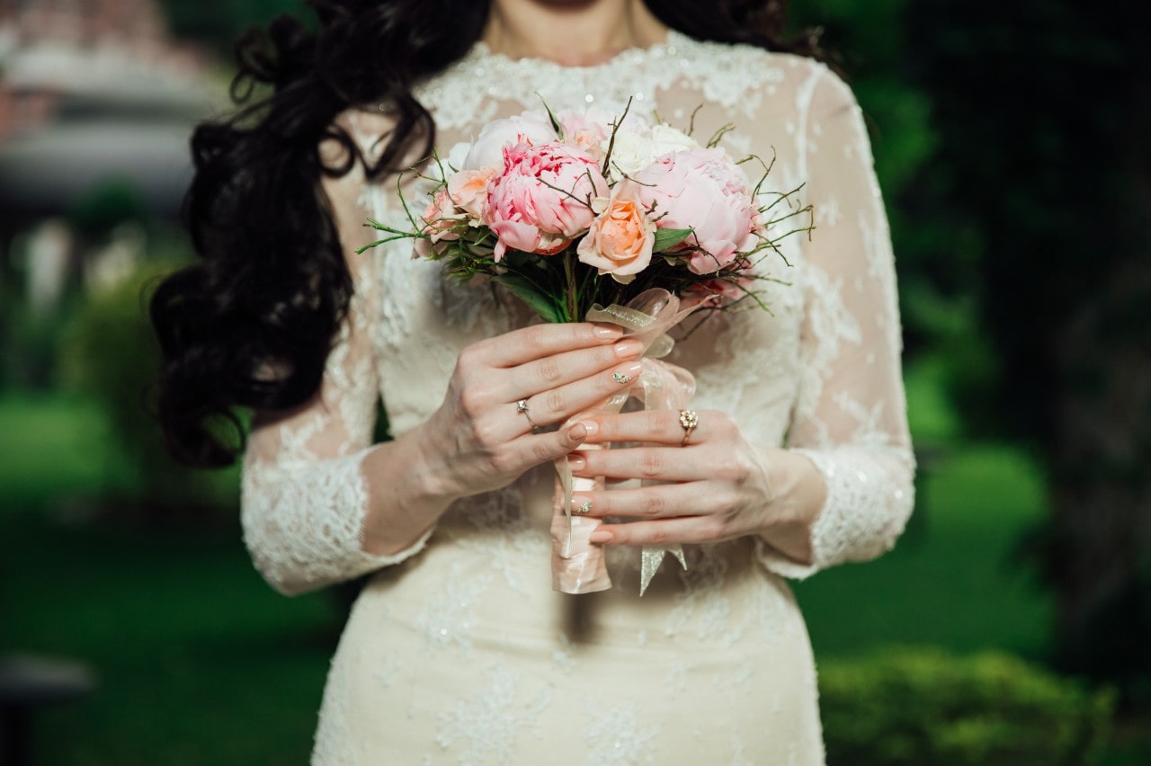 A close up of a bride with long black hair holding a bouquet with peonies, showcasing diamond solitaire engagement ring. A close up of a bride with long black hair holding a bouquet with peonies, showcasing diamond solitaire engagement ring.