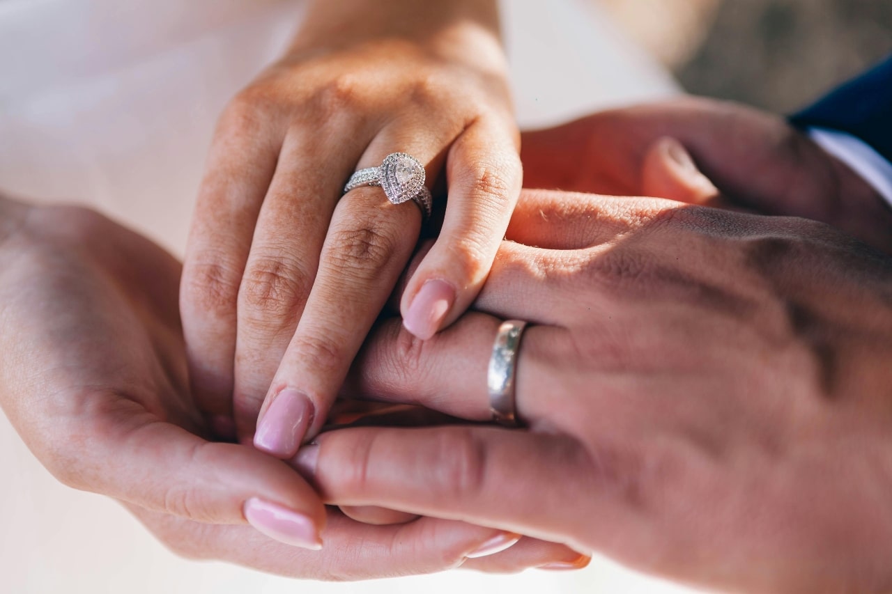 A close-up of a couple’s hands, with emphasis on his wedding band and the ornate pear cut halo engagement ring on her finger.