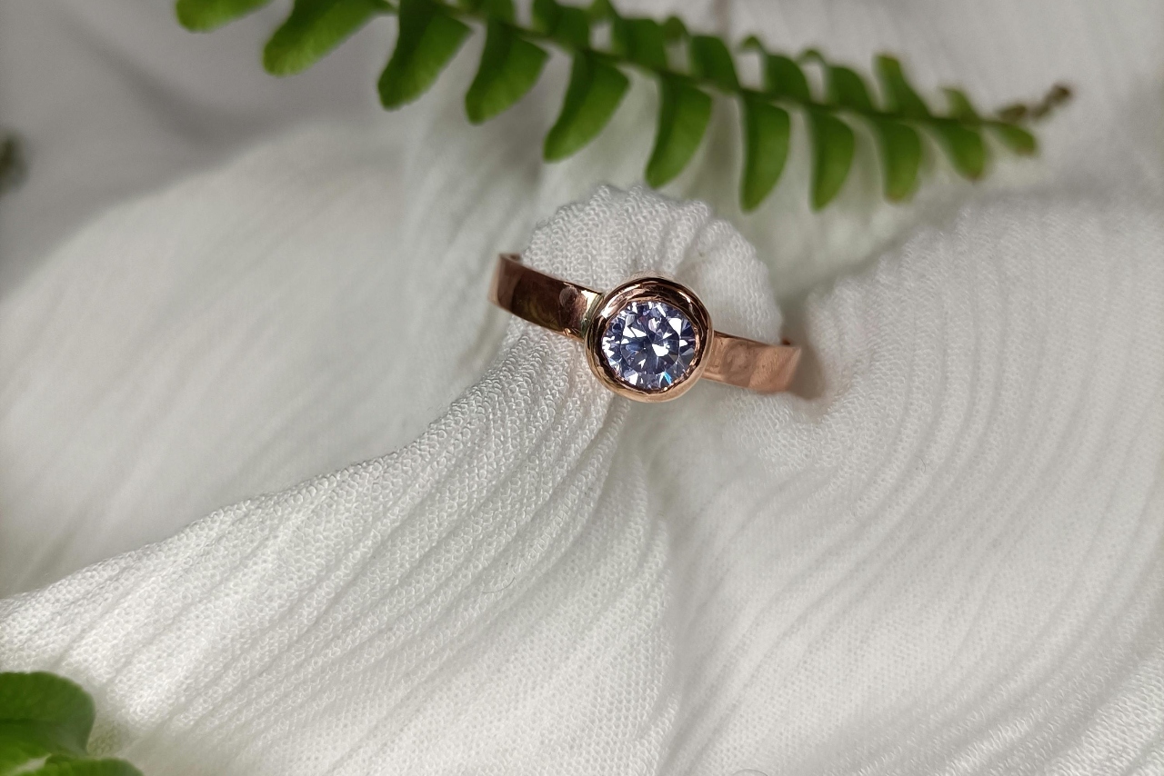 A close-up of a rose gold bezel engagement ring with a round cut diamond, laying on white fabric with some greenery draped around.
