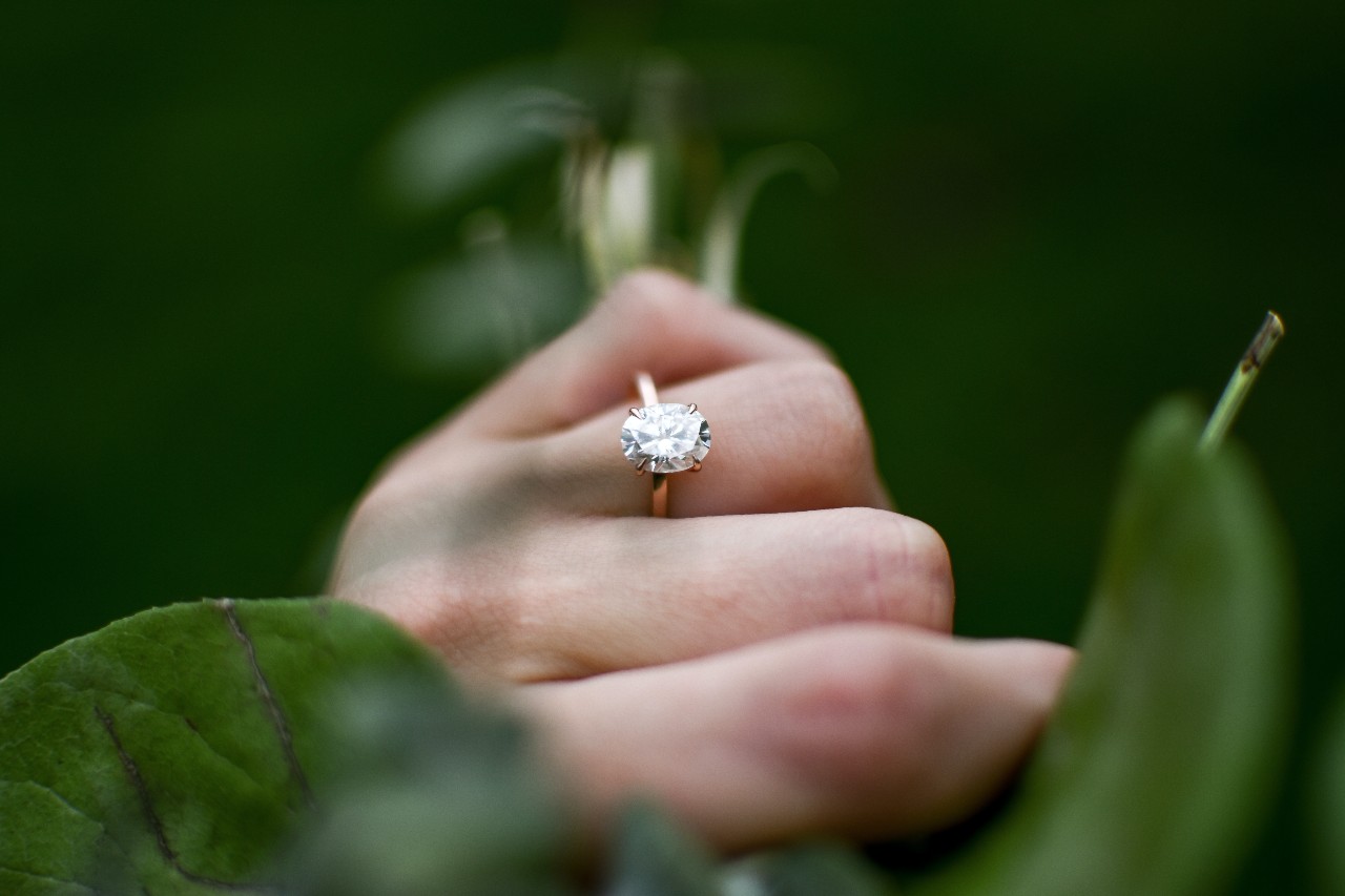 A close up image of a hand wearing a solitaire engagement ring, surrounded in greenery