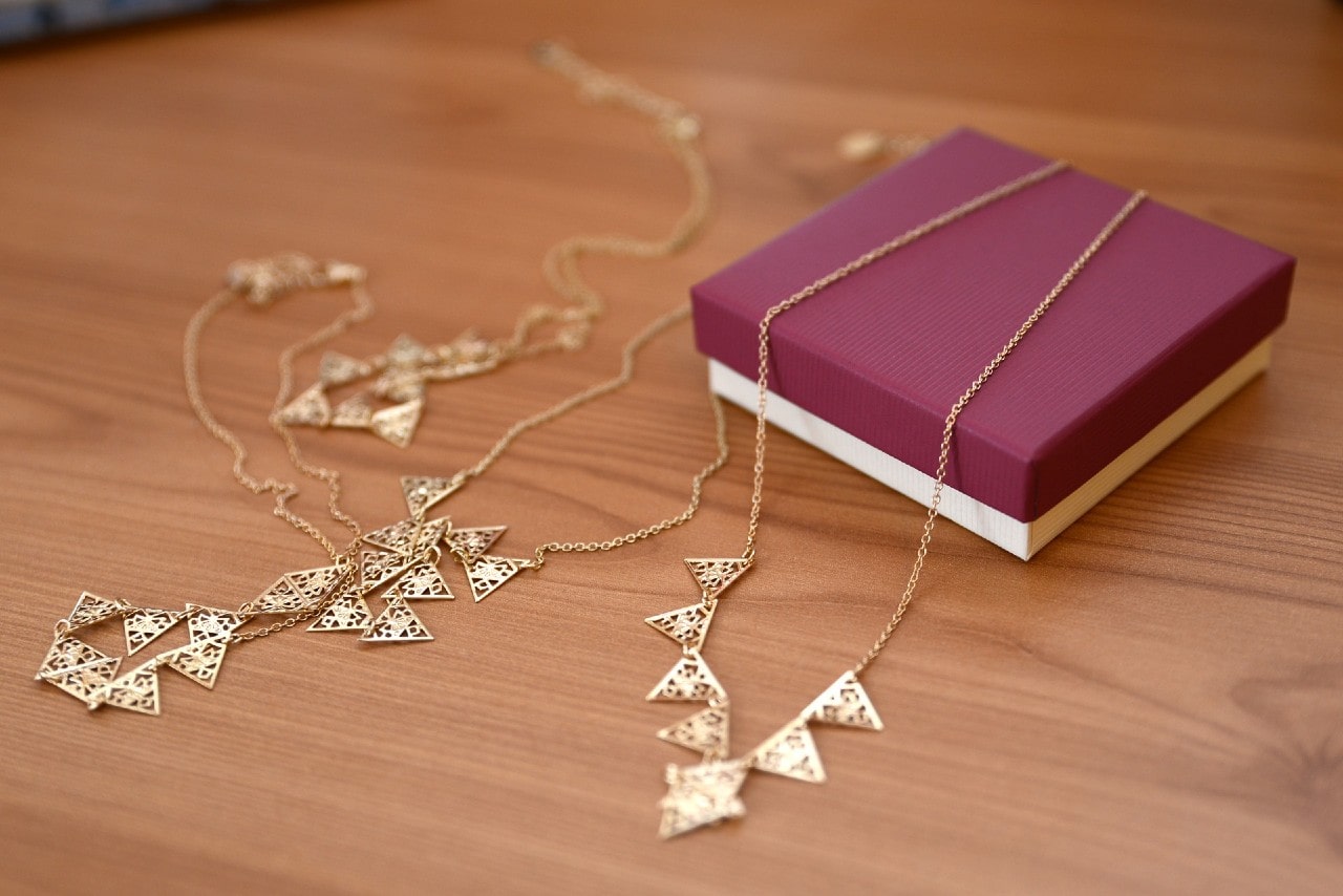 Two gold necklaces with triangular designs displayed beside a purple jewelry box on a wooden surface