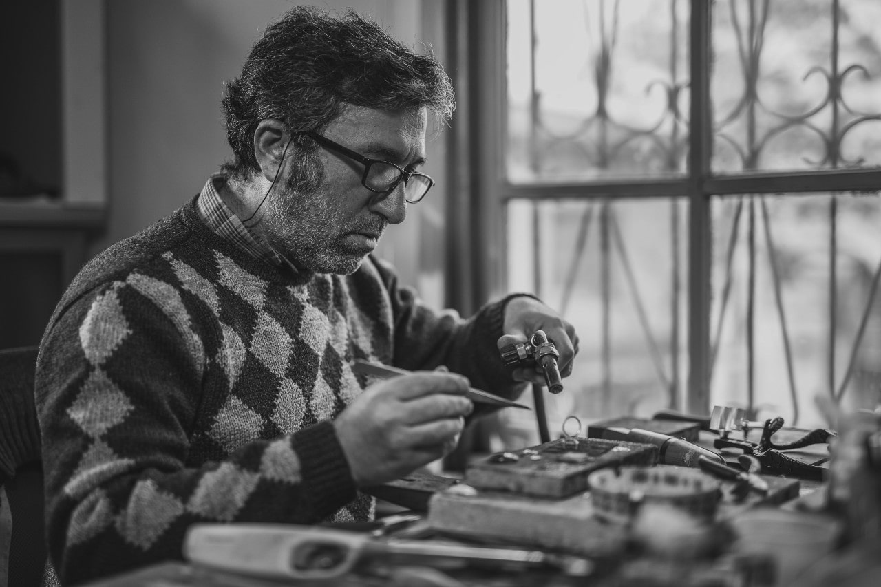 A black and white image of a jeweler servicing a large ring at a desk beside a window.