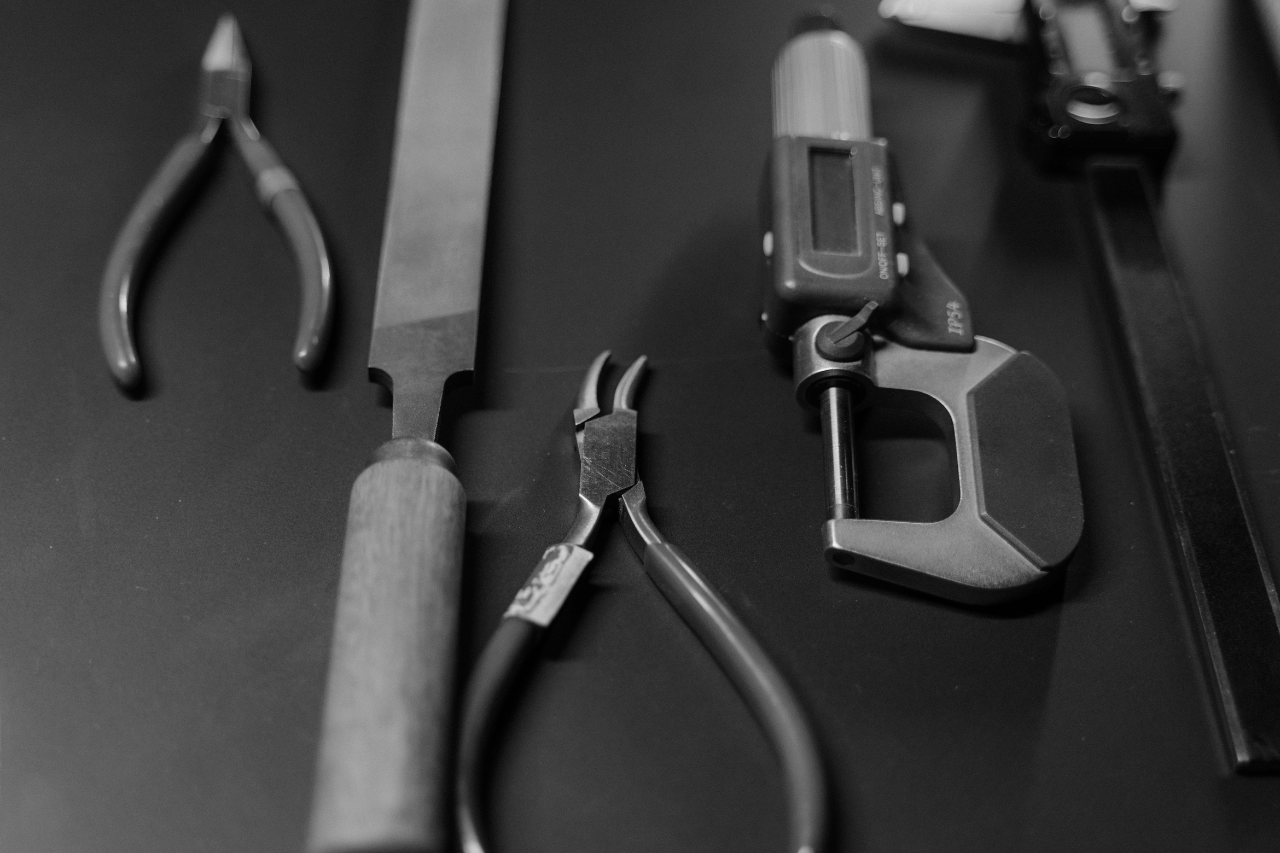 A black and white close-up of jewelers’ tools including pliers, a large file, and digital jewelry calipers atop a dark mat.